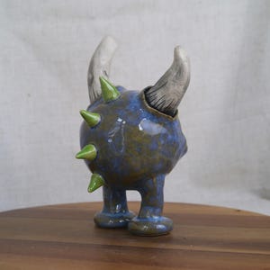 May include: A ceramic sculpture of a creature with a blue and green speckled body, two curved horns, and green spikes. The sculpture has two small legs and is displayed on a wooden surface.