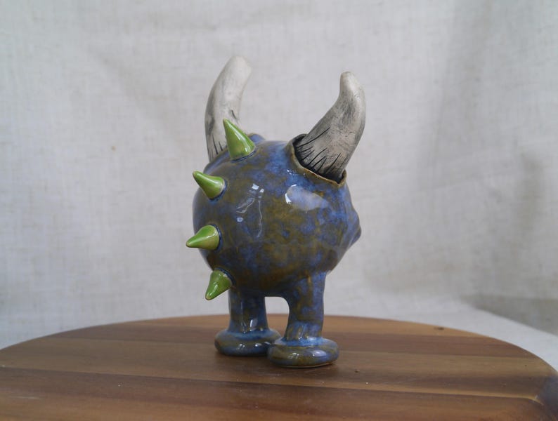 May include: A ceramic sculpture of a creature with a blue and green speckled body, two curved horns, and green spikes. The sculpture has two small legs and is displayed on a wooden surface.