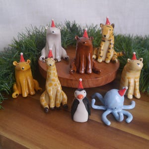 May include: A selection of charming animal ornaments, such as a bear, tiger, giraffe, penguin, and octopus, all adorned with red and white Santa hats. The figurines are displayed on a wooden surface with festive greenery.