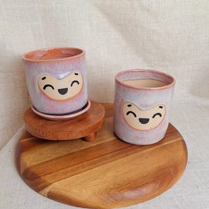May include: Two ceramic planters with a smiling face design. The planters are light purple with a peach-colored rim and a tan face. One planter sits on a small wooden stand with a matching saucer. The other planter sits on a wooden cutting board.