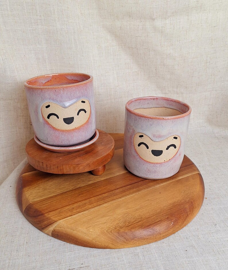 May include: Two ceramic planters with a smiling face design. The planters are light purple with a peach-colored rim and a tan face. One planter sits on a small wooden stand with a matching saucer. The other planter sits on a wooden cutting board.