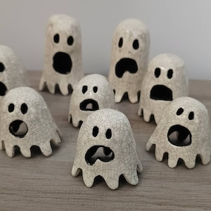 May include: A group of seven white ceramic ghost figurines with black eyes and mouths. The ghosts have a crackled glaze finish and are arranged on a wooden surface.