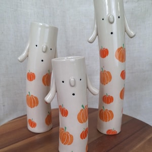 May include: Three white ceramic vases with a pumpkin pattern. The vases are shaped like people with arms and a face. The vases are different sizes.
