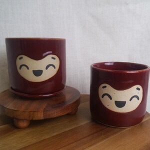 May include: Two maroon ceramic planters with a smiling face design. One planter sits on a wooden stand with a matching saucer. The other planter is smaller and sits on a wooden surface. The planters are cylindrical.