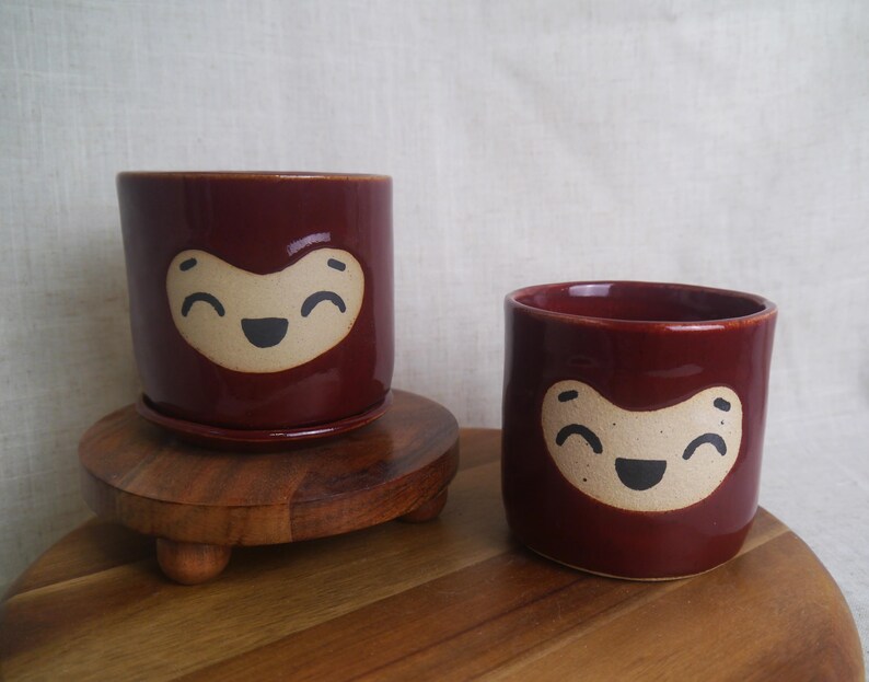 May include: Two maroon ceramic planters with a smiling face design. One planter sits on a wooden stand with a matching saucer. The other planter is smaller and sits on a wooden surface. The planters are cylindrical.
