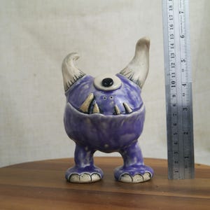 May include: A whimsical, handmade ceramic monster figurine. The purple creature has one eye, two horns, and a toothy grin. The figurine is approximately 4 inches tall and stands on two feet. The horns and teeth are off-white.