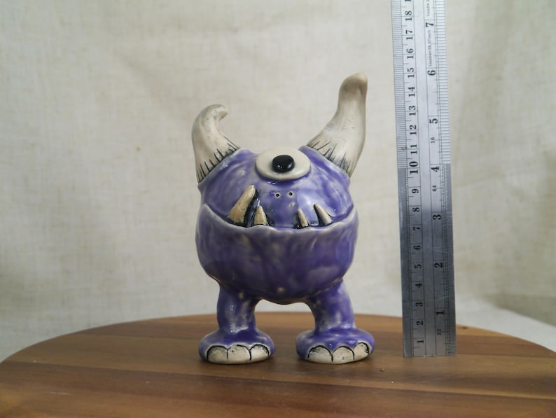 May include: A whimsical, handmade ceramic monster figurine. The purple creature has one eye, two horns, and a toothy grin. The figurine is approximately 4 inches tall and stands on two feet. The horns and teeth are off-white.
