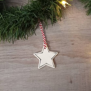 May include: A white star-shaped Christmas ornament with a black outline, hanging from a red and white twine. The ornament is set against a wooden background, with green garland and small yellow lights.