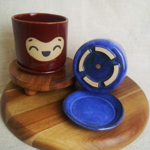May include: A ceramic planter with a smiling face design in a deep red color. The planter sits on a wooden stand, with a matching red saucer. A blue planter and saucer are also present.