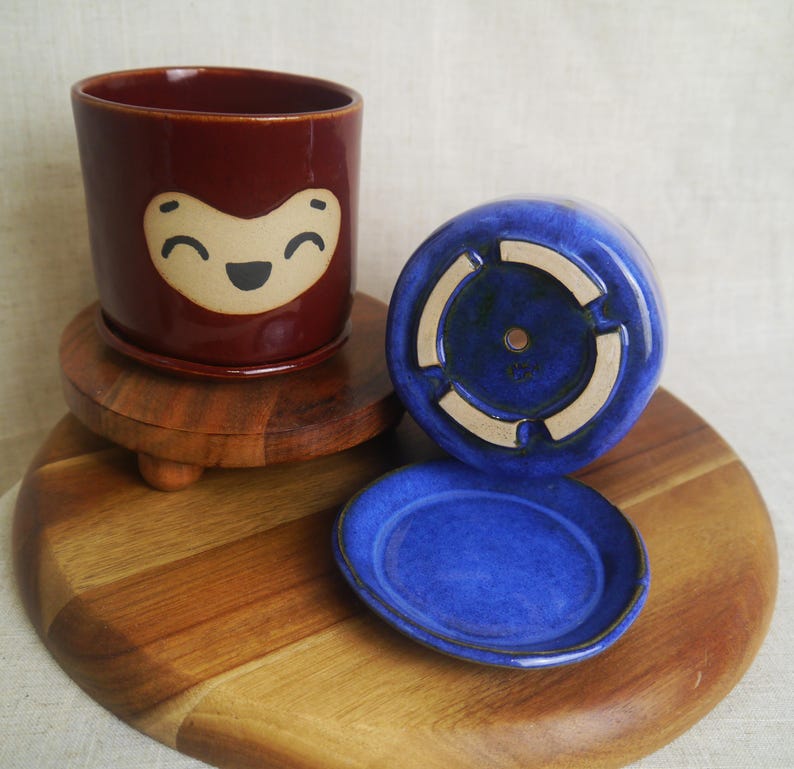 May include: A ceramic planter with a smiling face design in a deep red color. The planter sits on a wooden stand, with a matching red saucer. A blue planter and saucer are also present.