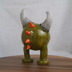 May include: A whimsical ceramic figurine of a creature with a rounded green body, orange spikes, and two curved white horns. The figurine has two short legs and is standing on a wooden surface.