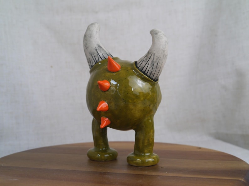 May include: A whimsical ceramic figurine of a creature with a rounded green body, orange spikes, and two curved white horns. The figurine has two short legs and is standing on a wooden surface.