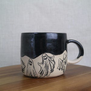 May include: A black and white ceramic mug with a black glaze and a white band featuring a repeating pattern of ghosts. The mug has a rounded handle.
