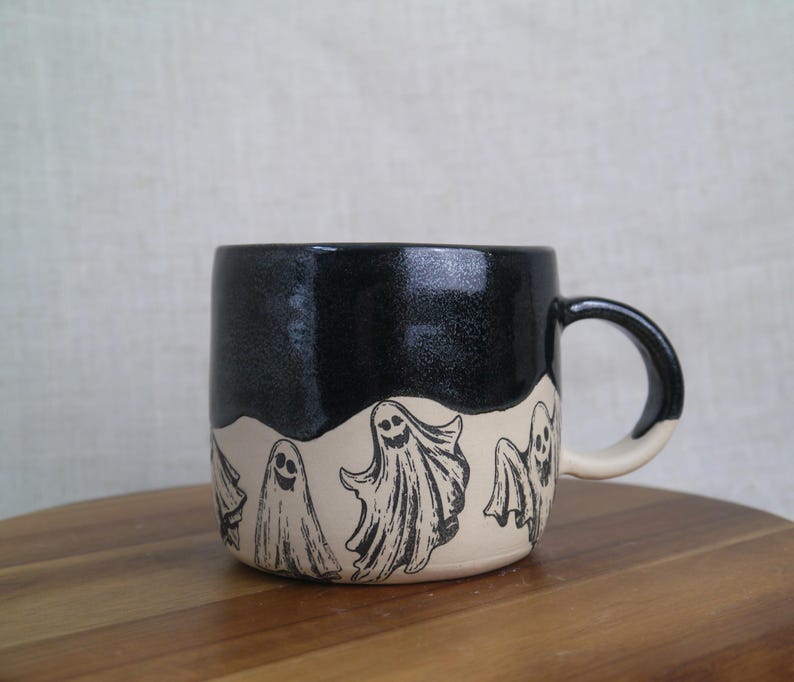 May include: A black and white ceramic mug with a black glaze and a white band featuring a repeating pattern of ghosts. The mug has a rounded handle.