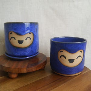 May include: Two blue ceramic planters with a smiling face design. One planter sits on a matching saucer, atop a wooden stand. The other planter is smaller and sits directly on a wooden surface. The planters are glazed with a glossy finish.