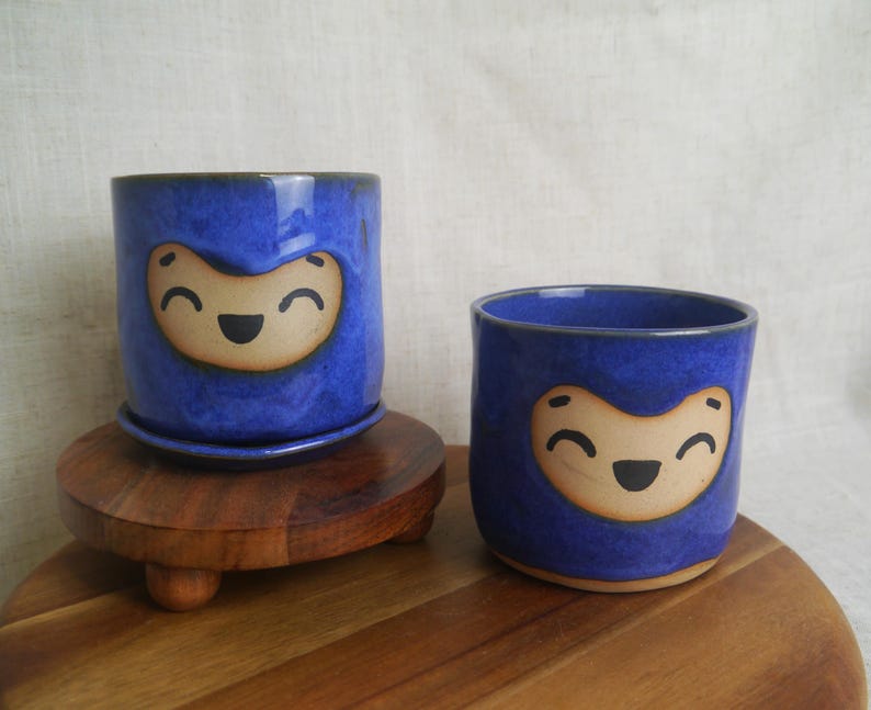 May include: Two blue ceramic planters with a smiling face design. One planter sits on a matching saucer, atop a wooden stand. The other planter is smaller and sits directly on a wooden surface. The planters are glazed with a glossy finish.