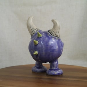 May include: A whimsical ceramic figurine in the shape of a creature with a purple body, two white horns, and green spikes. The figurine has two small feet and is set on a wooden surface.