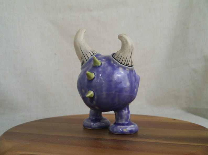 May include: A whimsical ceramic figurine in the shape of a creature with a purple body, two white horns, and green spikes. The figurine has two small feet and is set on a wooden surface.