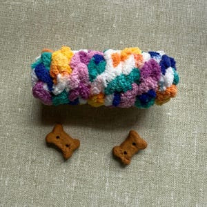 May include: A colorful, hand-knitted dog toy in shades of blue, green, orange, yellow, pink, and white. Two bone-shaped dog biscuits are next to the toy. The background is a light green fabric.