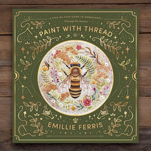 May include: A square book cover with the title "Paint With Thread" and the author's name, Emillie Ferris. The cover is dark green with gold accents and illustrations of flowers, stars, and a bee. The book is a step-by-step guide to embroidery.