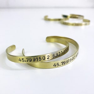 Könnte beinhalten: Zwei goldene Metallarmbänder mit den Zahlen "45.7991502 6.9218539" eingraviert.