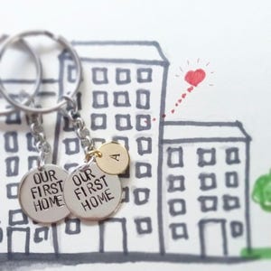 Könnte beinhalten: Ein silberner Schlüsselanhänger mit zwei runden Anhängern, auf denen "Our First Home" steht, und einem goldenen Anhänger mit dem Buchstaben "A". Der Schlüsselanhänger hängt an einer Zeichnung eines Gebäudes mit einem roten Herzen darüber.