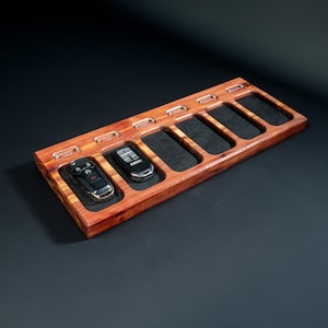May include: A rectangular wooden key holder with six slots, each lined with black fabric. Two car keys are placed in the holder. The wood has a reddish-brown hue, and the design includes clear acrylic accents.