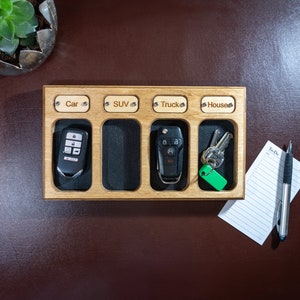 Puede incluir: Organizador de llaves de madera con cuatro ranuras etiquetadas: Coche, SUV, Camión y Casa. Cada ranura contiene un juego de llaves o un llavero. El organizador está sobre una superficie marrón, junto a un bloc de notas y un bolígrafo.