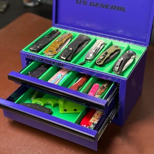 Puede incluir: Un cofre de herramientas azul US General con tres cajones, cada uno con varias navajas plegables. El interior es verde, lo que contrasta con el exterior azul. La parte superior del cofre está abierta, revelando más cuchillos.