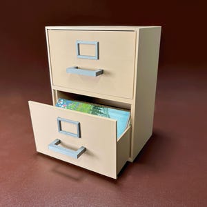May include: A small, two-drawer filing cabinet in a light beige color. The top drawer is closed, while the bottom drawer is open, revealing colorful artwork. The cabinet has gray handles and label holders.