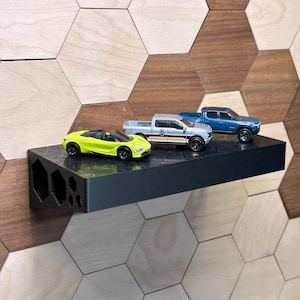 May include: Three toy cars on a black shelf with a honeycomb pattern background. The cars are a yellow sports car, a silver pickup truck, and a blue pickup truck.
