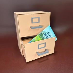 May include: A small, two-drawer filing cabinet in a light tan color. The top drawer is closed, while the bottom drawer is partially open, revealing a notebook with the words "Field Notes" on the cover. The cabinet has gray handles and label holders.