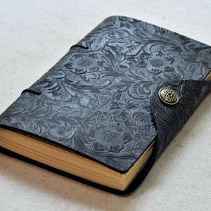 Pu&ograve; includere: Un diario in pelle nera con un design a rilievo floreale e una chiusura a bottone in ottone.