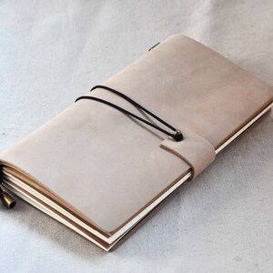 May include: A light brown leather bound journal with a black elastic closure and brass accents. The journal is closed and lying flat on a light beige fabric surface.