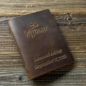 May include: A brown leather-bound journal with the text "The Officiant" embossed on the front cover. The names "John and Ashley" and the date "September 16, 2023" are also embossed on the cover.