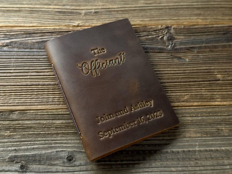 May include: A brown leather-bound journal with the text "The Officiant" embossed on the front cover. The names "John and Ashley" and the date "September 16, 2023" are also embossed on the cover.
