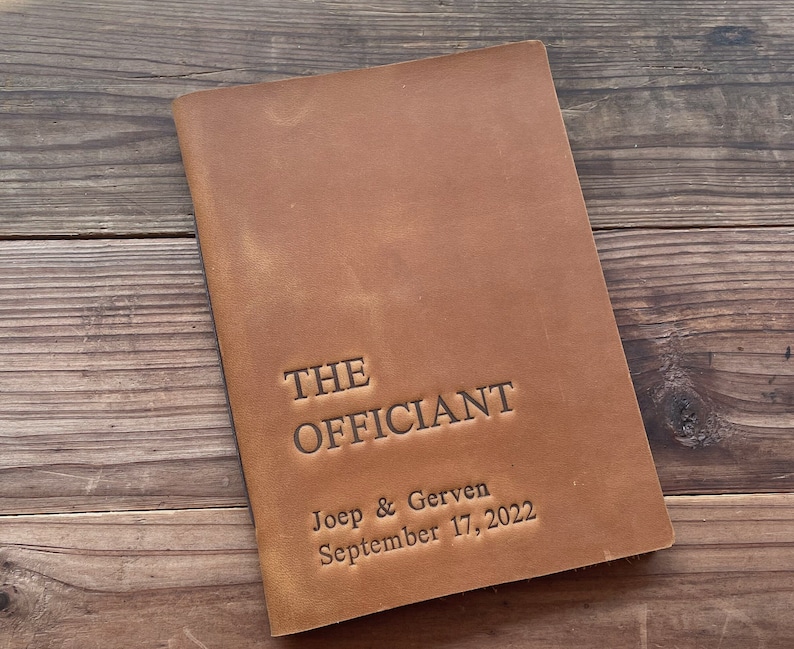 May include: A brown leather-bound journal with the text "THE OFFICIANT" embossed on the front cover. The text "Joep & Gerven September 17, 2022" is also embossed on the cover.