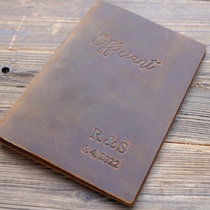 May include: A brown leather-bound journal with the word "Officiant" embossed on the front cover. The initials "R & S" and the date "6.4.2022" are also embossed on the cover.
