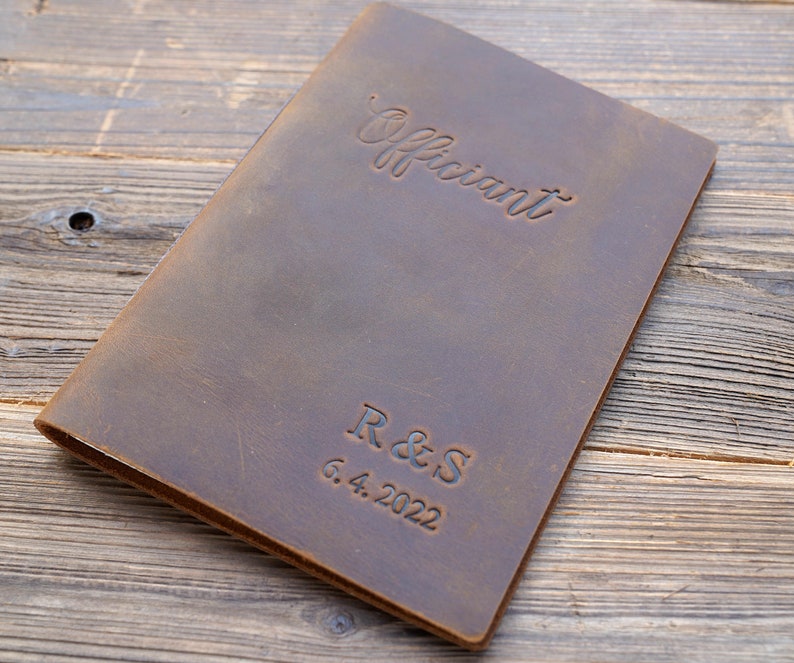May include: A brown leather-bound journal with the word "Officiant" embossed on the front cover. The initials "R & S" and the date "6.4.2022" are also embossed on the cover.