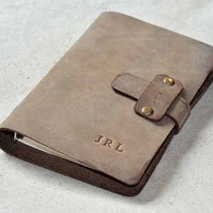 May include: A brown leather journal with a strap closure and brass accents. The journal has the initials "JRL" embossed on the front cover.
