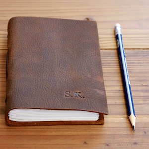 May include: A brown leather-bound journal with the initials "S.K." embossed on the front cover. A pencil is lying next to the journal on a wooden surface.