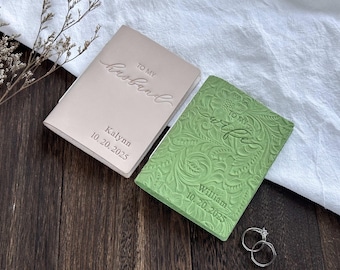 May include: Two personalized wedding day journals, one blush pink and one green, with embossed text. The pink journal reads "TO MY Husband" and the green journal reads "TO MY Wife". Both journals have names and dates. Two silver rings are also visible.