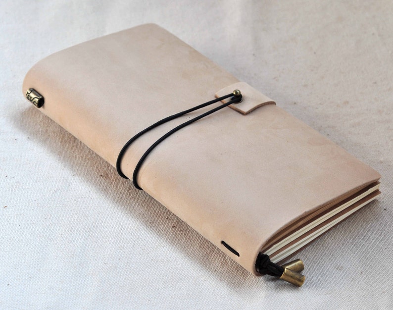 May include: A light brown leather bound journal with a black elastic closure and brass accents. The journal is closed and lying on a white fabric surface.