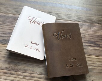 May include: Two brown leather vow books with the word "Vows" embossed on the front. The book on the left is white and has the names "Kelly" and the date "10. 9. 2021" embossed on the front. The book on the right has the name "Alex" and the date "8. 29. 2021" embossed on the front.