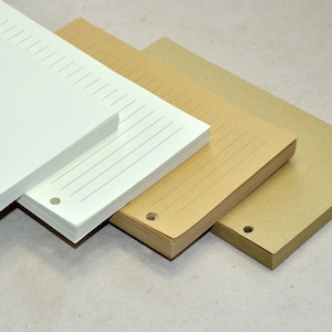 Pu&ograve; includere: Tre pile di carta rigata, una bianca, una marrone e una beige. Ogni pila ha tre fori perforati sul lato sinistro.