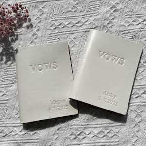 May include: Two off-white vow books with the word "VOWS" embossed on the cover. One book is inscribed with "Megan 9.7.2025" and the other with "Ricky 9.7.2025". The books are on a white textured surface.