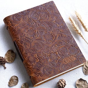 Può includere: Un diario in pelle marrone con un design floreale in rilievo. La copertina presenta un motivo dettagliato di fiori e foglie. Il diario è chiuso con un cinturino in pelle. Le iniziali "GW" sono visibili.
