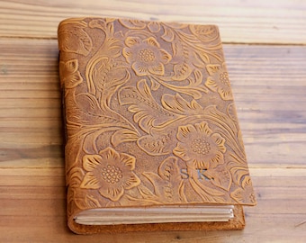 May include: A brown leather journal with a floral embossed design. The journal has a spine with multiple pages and the initials "S.K." are embossed on the front cover.