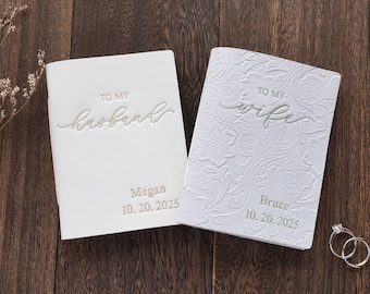 May include: Two white wedding books, one embossed with floral patterns and the other with a smooth finish. The first book reads "To My Husband" and the second reads "To My Wife". Both books have names and dates. Wedding rings are in the corner.