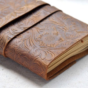 May include: A brown leather journal with a floral embossed design. The journal is closed and has a leather strap wrapped around it.